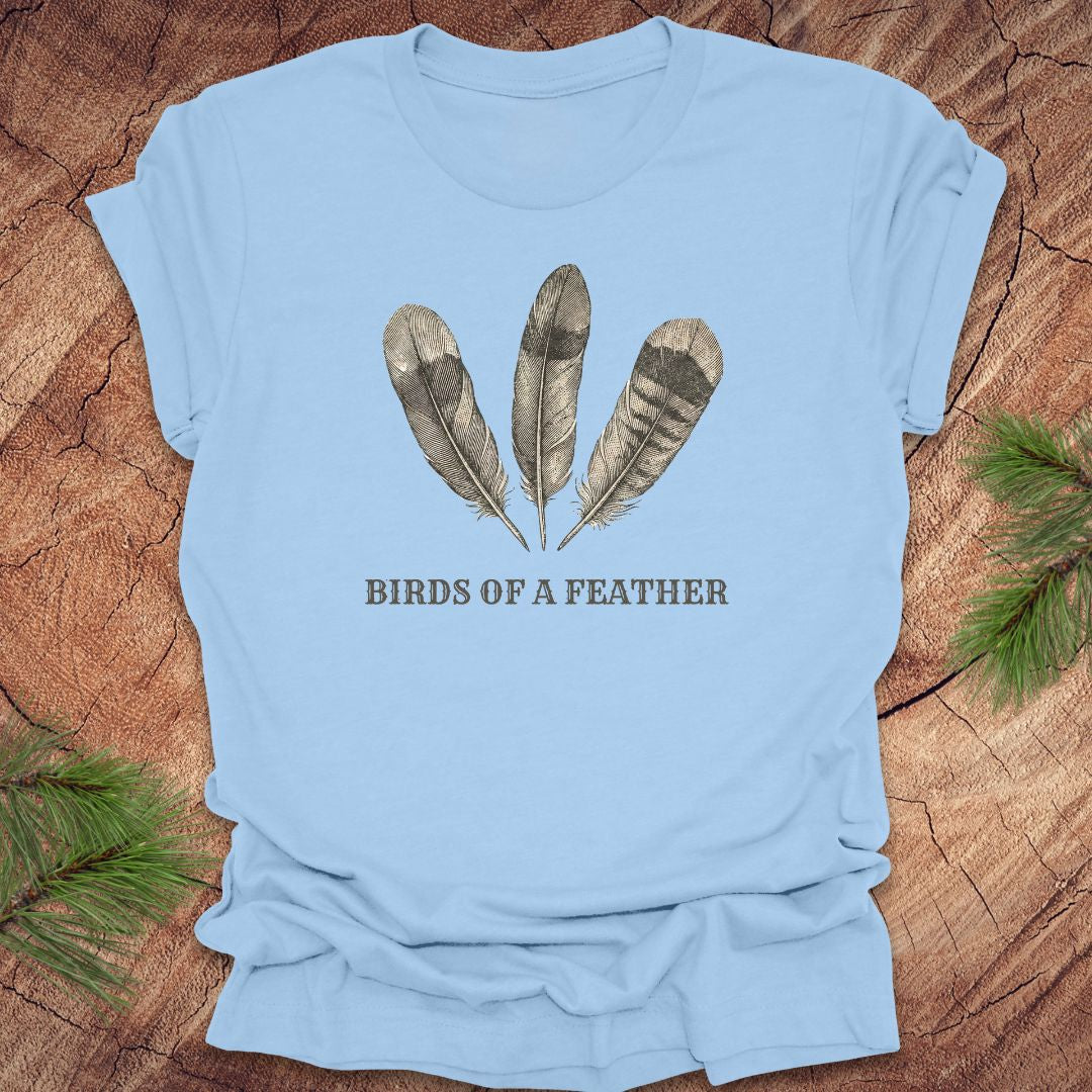 Light blue t-shirt with feather design and 'Birds of a Feather' text on a wooden surface.