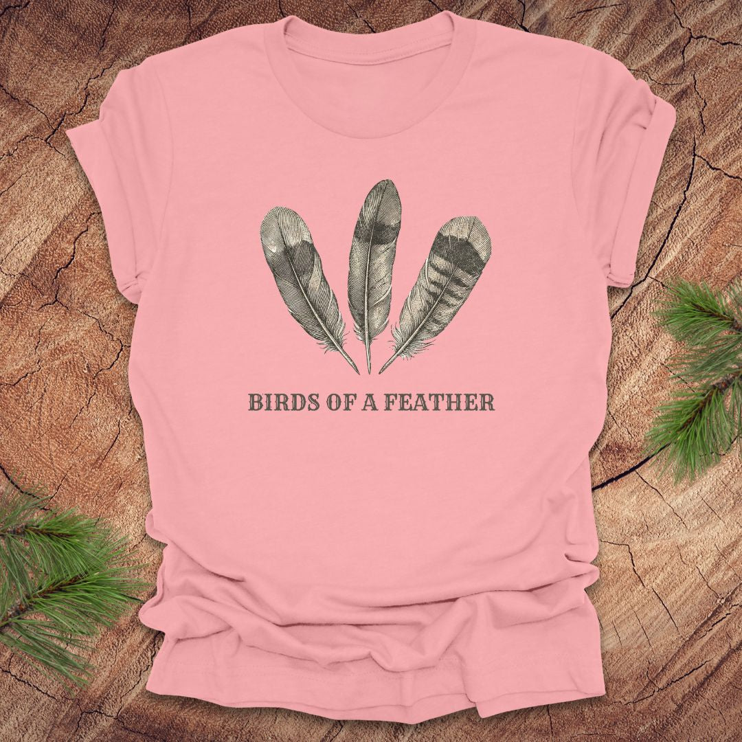 Pink t-shirt with feather design and 'Birds of a Feather' text on a wooden surface.