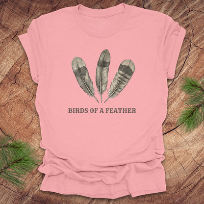 Pink t-shirt with feather design and 'Birds of a Feather' text on a wooden surface.