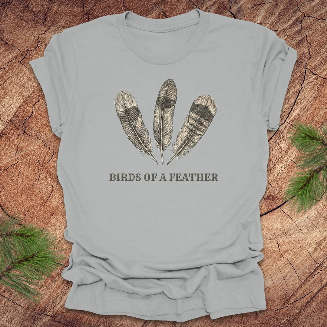 Light gray t-shirt with feather design and 'Birds of a Feather' text on a wooden surface.