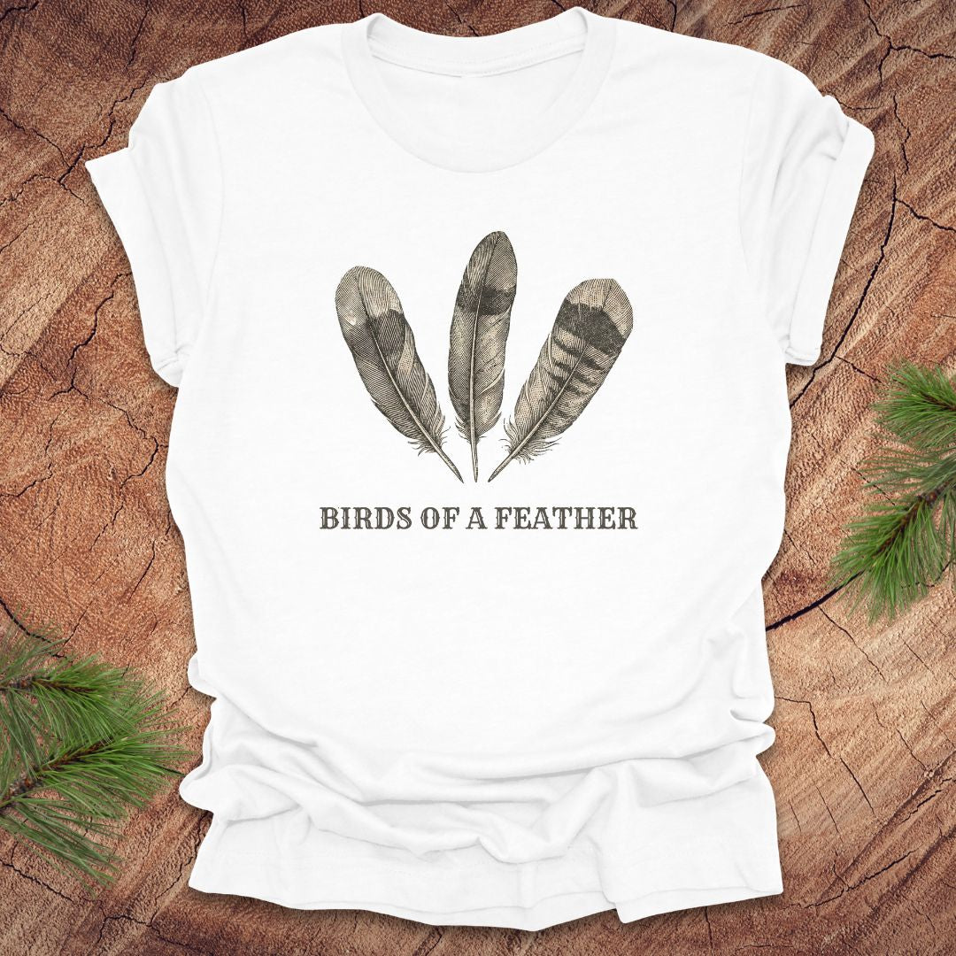 White t-shirt with feather design and 'Birds of a Feather' text on a wooden surface.