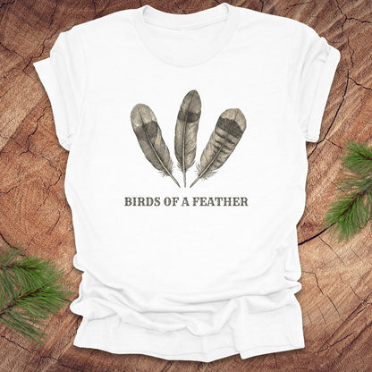 White t-shirt with feather design and 'Birds of a Feather' text on a wooden surface.