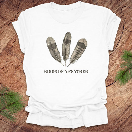 White t-shirt with feather design and 'Birds of a Feather' text on a wooden surface.