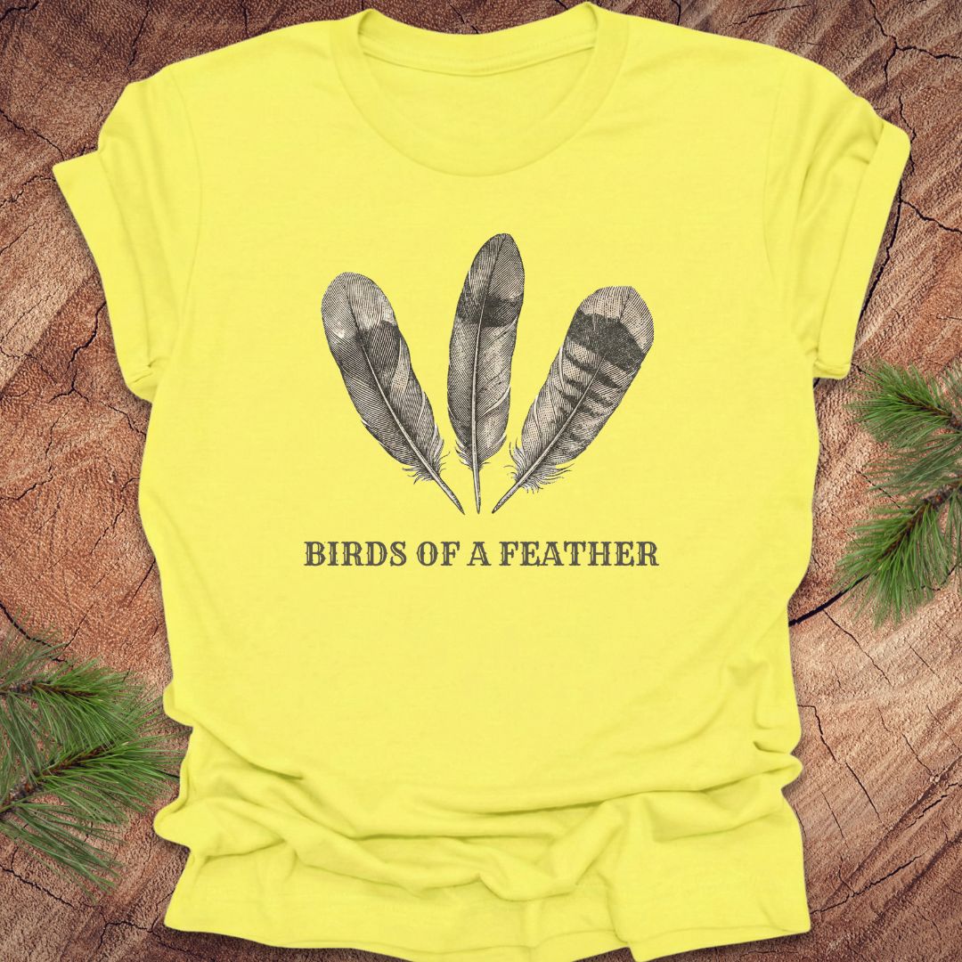 Yellow t-shirt with feather design and 'Birds of a Feather' text on a wooden surface.