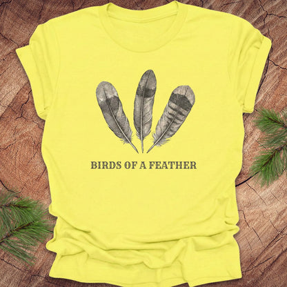 Yellow t-shirt with feather design and 'Birds of a Feather' text on a wooden surface.
