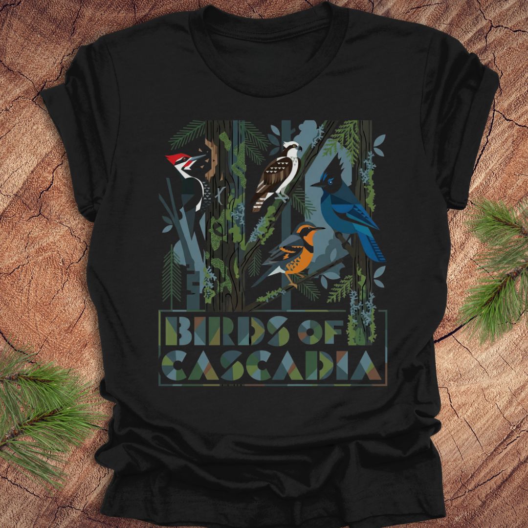 Black t-shirt with 'Birds of Cascadia' design on a wooden surface with pine needles.