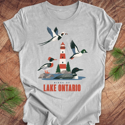 A light gray short-sleeve t-shirt with a graphic print of birds and a lighthouse on Lake Ontario, displayed flat on a rustic wood background with pine needles.