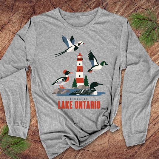 A heather gray long-sleeve t-shirt with a colorful graphic of birds and a lighthouse around Lake Ontario, displayed flat on a rustic wood background with pine needles.
