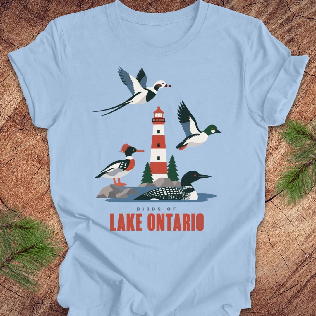 A light blue short-sleeve t-shirt laid flat on a rustic wood background with pine branches at the corners. The shirt features a colorful graphic of birds and a lighthouse with the text 'BIRDS OF LAKE ONTARIO' in red.