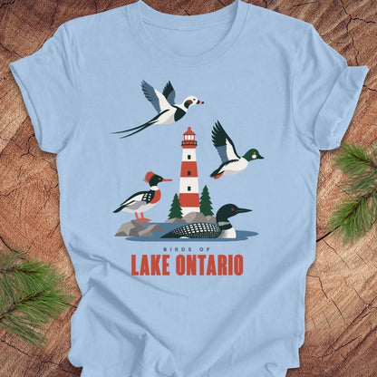 A light blue short-sleeve t-shirt laid flat on a rustic wood background with pine branches at the corners. The shirt features a colorful graphic of birds and a lighthouse with the text 'BIRDS OF LAKE ONTARIO' in red.