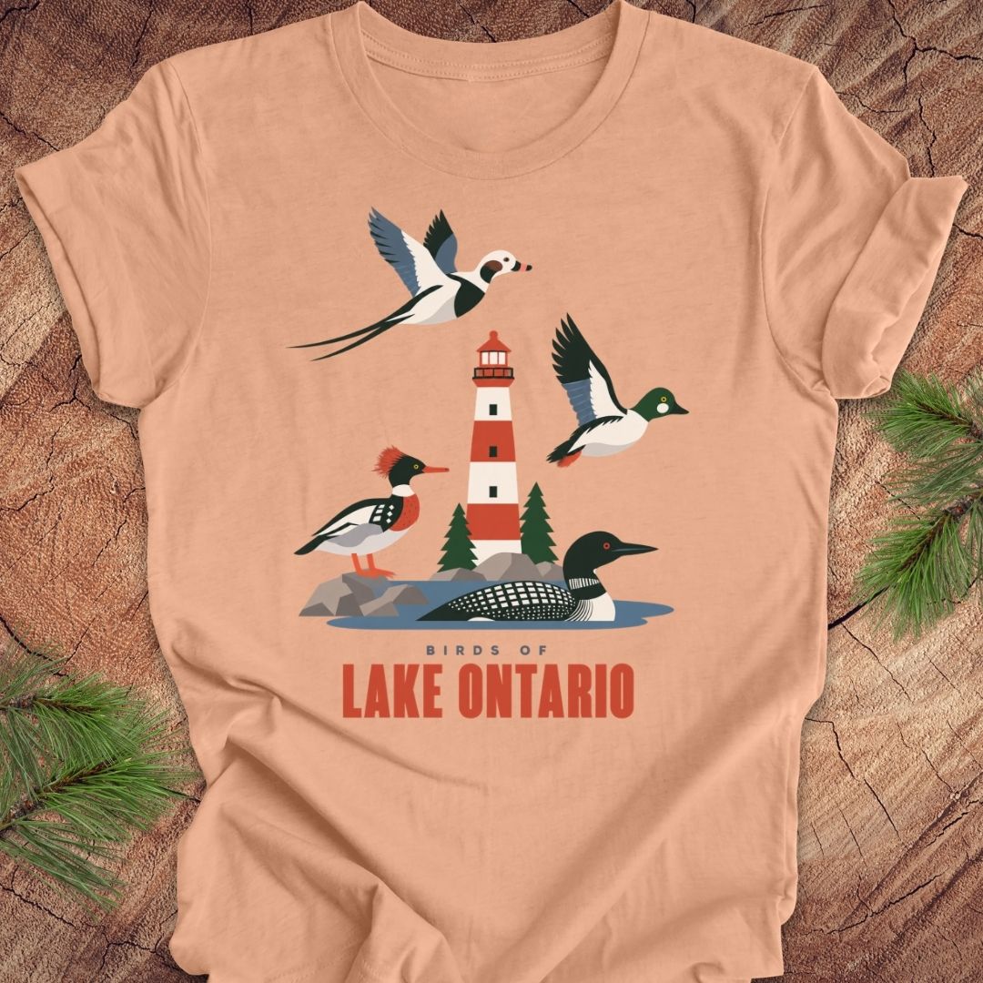 A peach-colored short-sleeve t-shirt with a graphic print of birds and a lighthouse on Lake Ontario, displayed flat on a rustic wood background with pine needles.