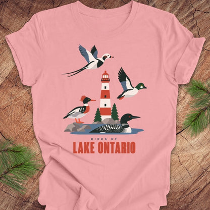 A pink short-sleeve crewneck t-shirt featuring a graphic print of birds and a lighthouse on Lake Ontario, styled flat on a rustic wood background with pine needles.