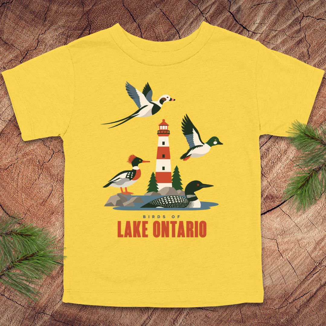 A yellow short-sleeve t-shirt with a graphic of birds and a lighthouse over Lake Ontario, displayed flat on a rustic wood background with pine branches.