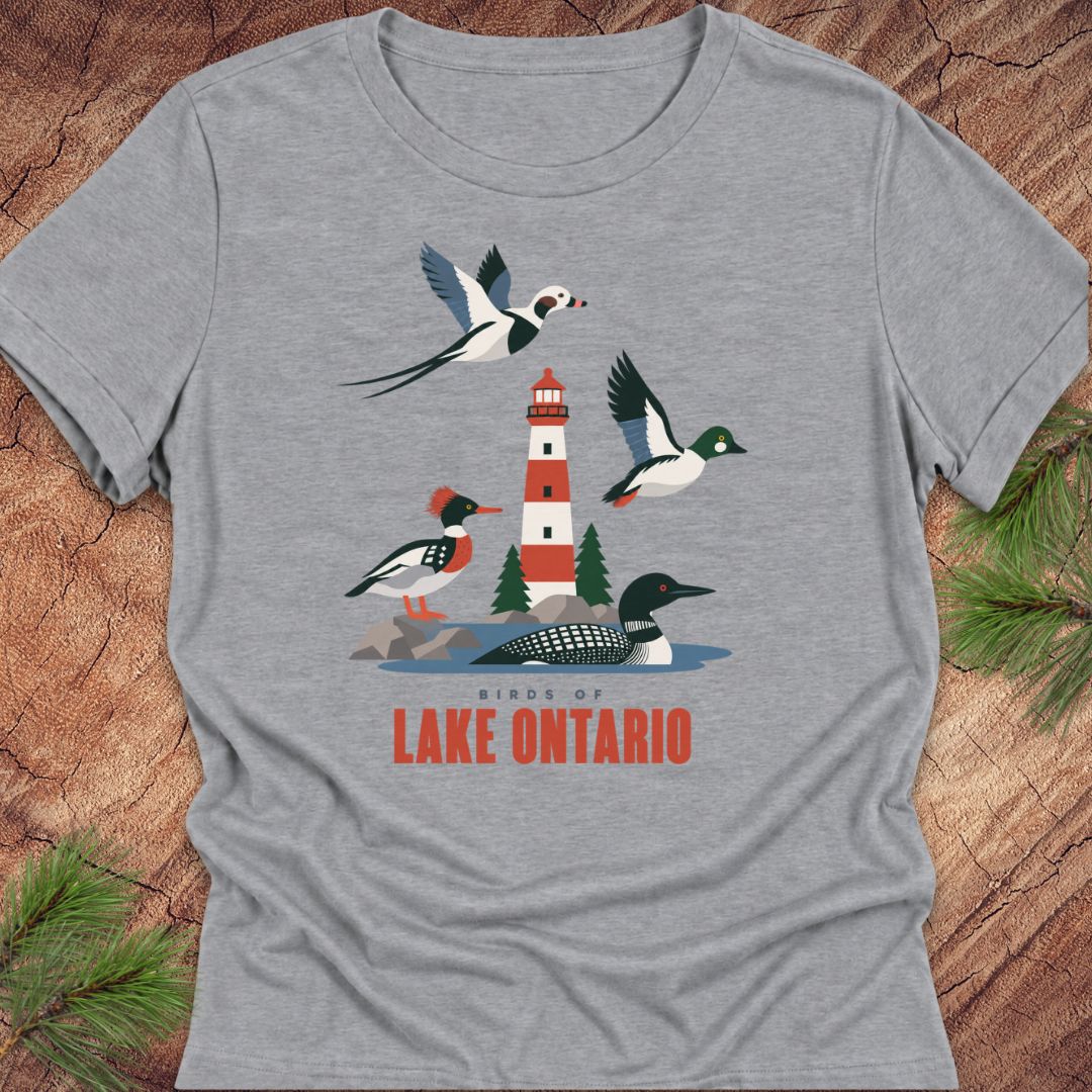 Athletic heather gray women's relaxed t-shirt with a red-and-white lighthouse, loon, merganser, goldeneye, and long-tailed duck above 'Birds of Lake Ontario' text, laid flat on a rustic wood background with pine branches.