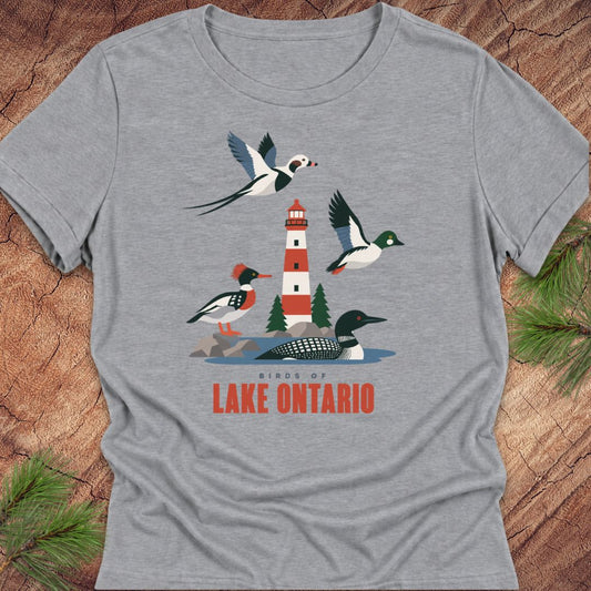 Athletic heather gray women's relaxed t-shirt with a red-and-white lighthouse, loon, merganser, goldeneye, and long-tailed duck above 'Birds of Lake Ontario' text, laid flat on a rustic wood background with pine branches.