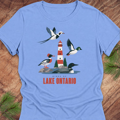 Carolina blue women's relaxed t-shirt with a red-and-white lighthouse, loon, merganser, goldeneye, and long-tailed duck above 'Birds of Lake Ontario' text, laid flat on a rustic wood background with pine branches.
