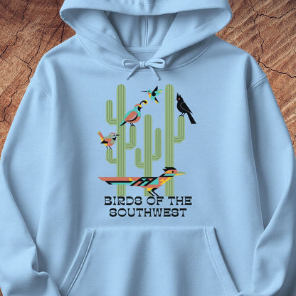 A light blue pullover hoodie featuring a colorful graphic of cacti and birds with the text 'BIRDS OF THE SOUTHWEST' printed on the front, displayed flat against a textured wood background.