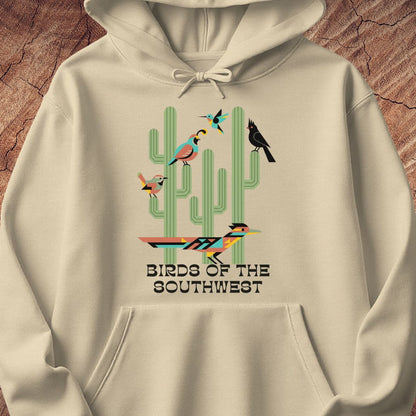 A cream-colored pullover hoodie featuring a graphic print of colorful birds perched on green cacti with the text 'BIRDS OF THE SOUTHWEST'. The hoodie is displayed flat against a textured brown wood background.
