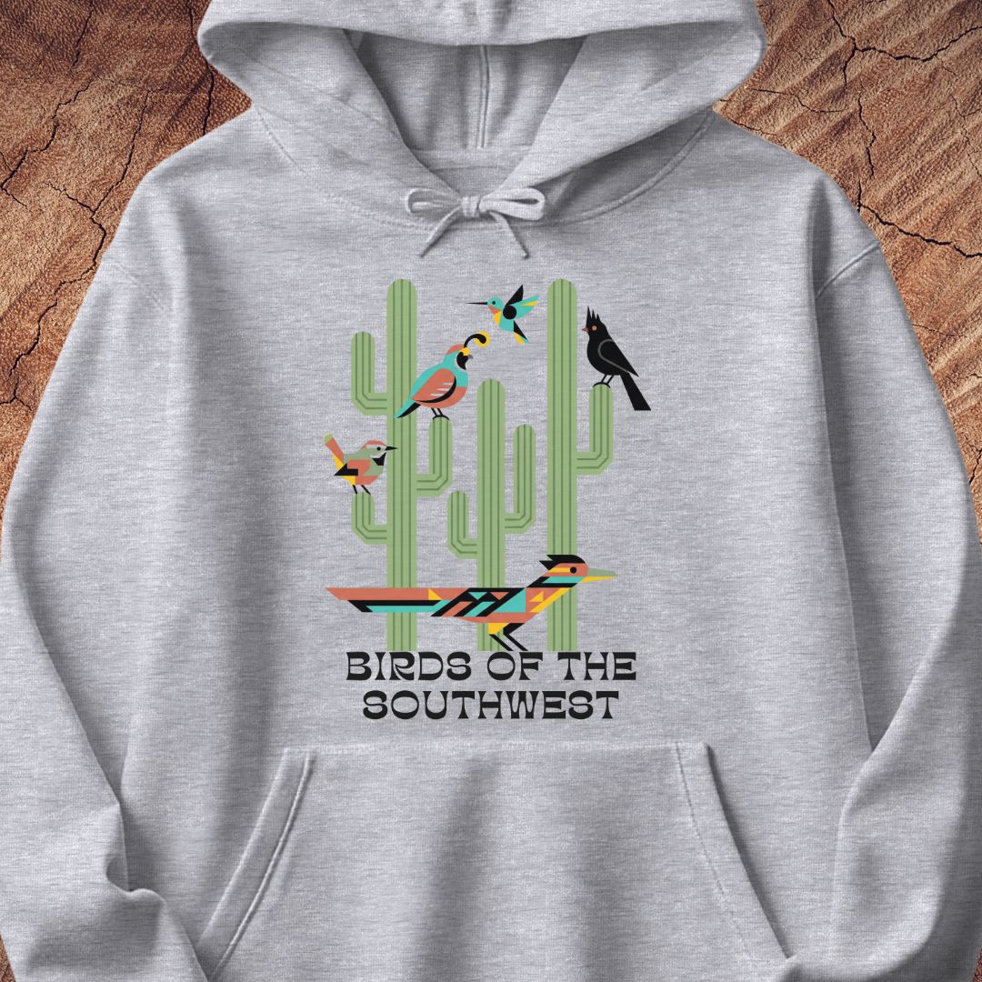 A light heather gray pullover hoodie featuring a colorful graphic of cacti and birds with the text 'BIRDS OF THE SOUTHWEST' printed on the front, displayed flat against a rustic wood background.