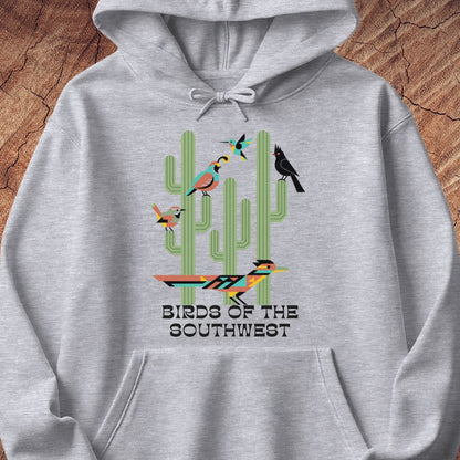 A light heather gray pullover hoodie featuring a colorful graphic of cacti and birds with the text 'BIRDS OF THE SOUTHWEST' printed on the front, displayed flat against a rustic wood background.