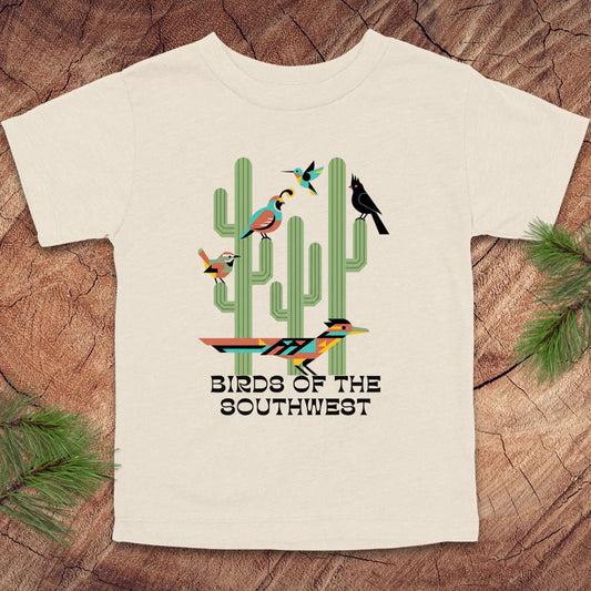 Beige toddler t-shirt with 'Birds of the Southwest' design featuring cacti and birds on a wooden surface.