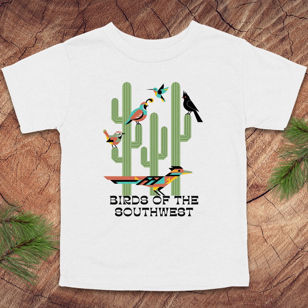 White toddler t-shirt with 'Birds of the Southwest' design featuring cacti and birds on a wooden surface.