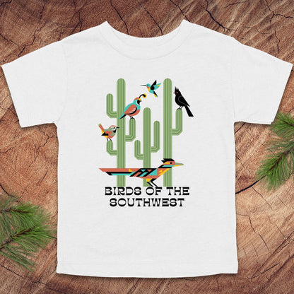 White toddler t-shirt with 'Birds of the Southwest' design featuring cacti and birds on a wooden surface.