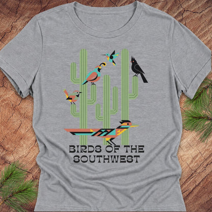 A heather gray short-sleeve t-shirt with a colorful graphic of stylized birds perched on green cacti and the text 'BIRDS OF THE SOUTHWEST'. The shirt is displayed flat on a rustic wood background with pine needles in the corners.