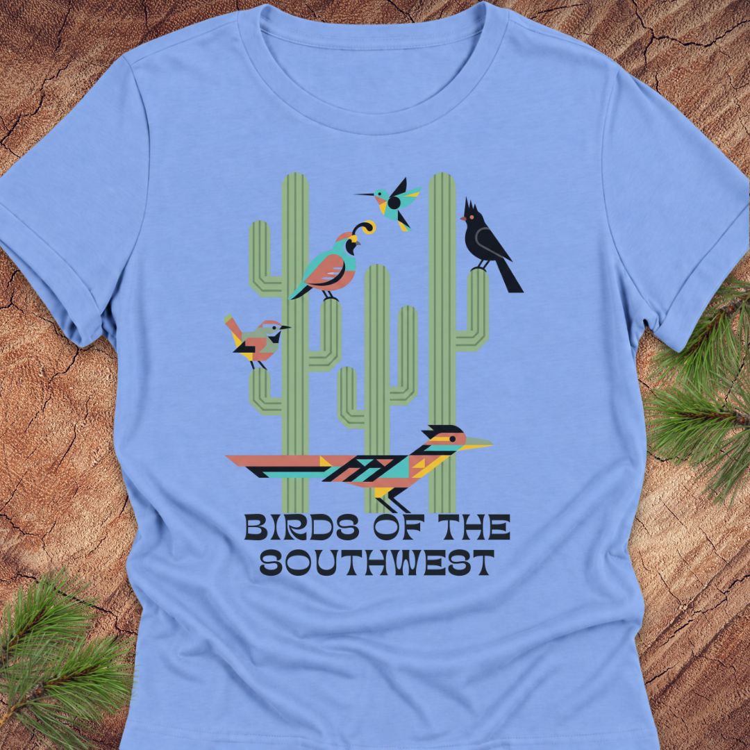 A light blue short-sleeve t-shirt laid flat on a rustic wood background with pine branches at the corners. The shirt features a colorful graphic of four stylized birds perched on green cacti, with the text 'BIRDS OF THE SOUTHWEST' printed below.