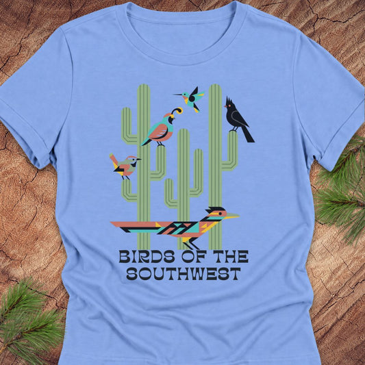A light blue short-sleeve t-shirt laid flat on a rustic wood background with pine branches at the corners. The shirt features a colorful graphic of four stylized birds perched on green cacti, with the text 'BIRDS OF THE SOUTHWEST' printed below.