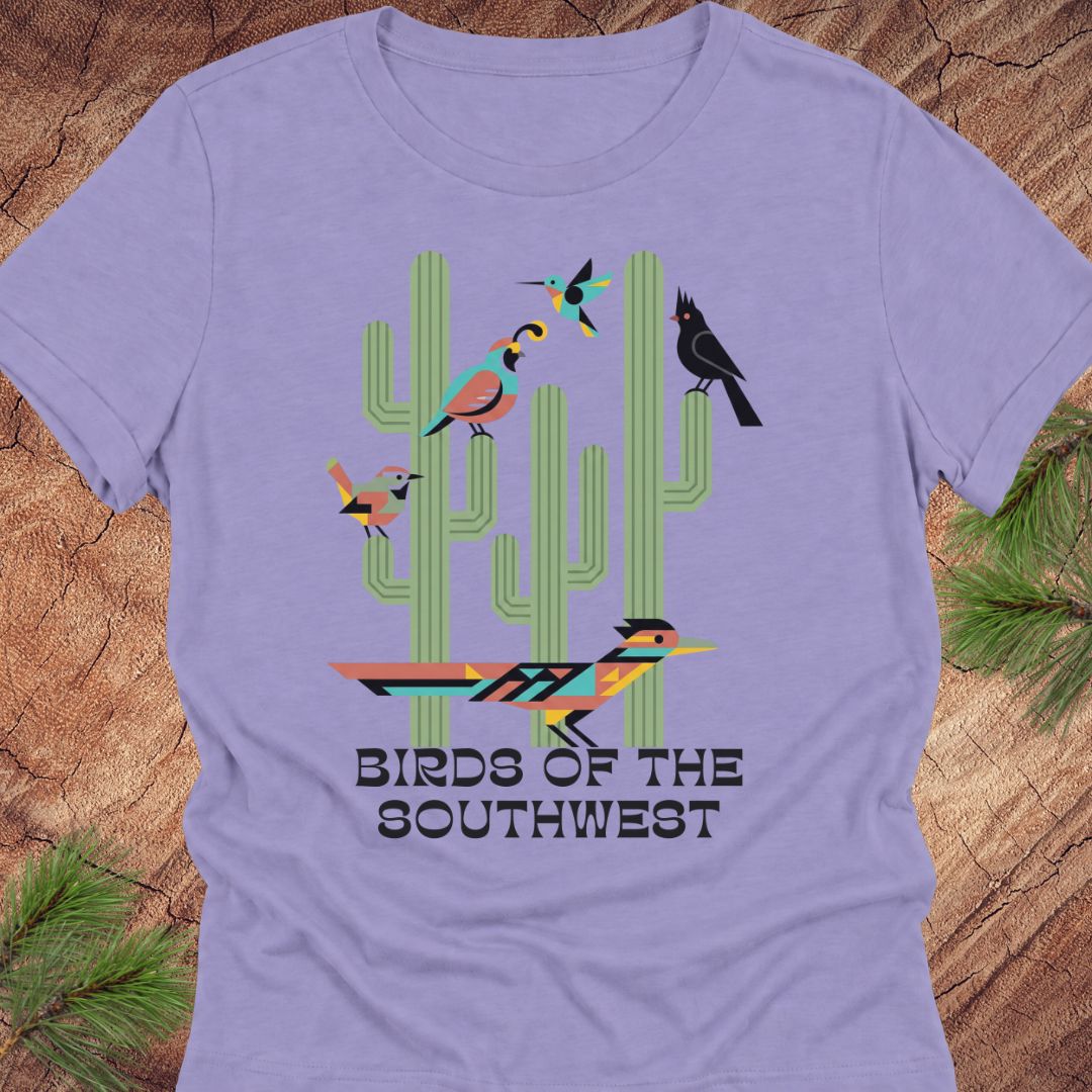 A lavender short-sleeve crewneck t-shirt featuring a colorful graphic of stylized birds perched on green cacti, with the text 'BIRDS OF THE SOUTHWEST' printed below. The shirt is displayed flat on a rustic wood background with pine needles in the corners.