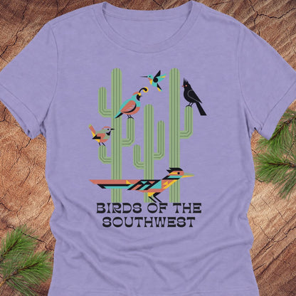 A lavender short-sleeve crewneck t-shirt featuring a colorful graphic of stylized birds perched on green cacti, with the text 'BIRDS OF THE SOUTHWEST' printed below. The shirt is displayed flat on a rustic wood background with pine needles in the corners.