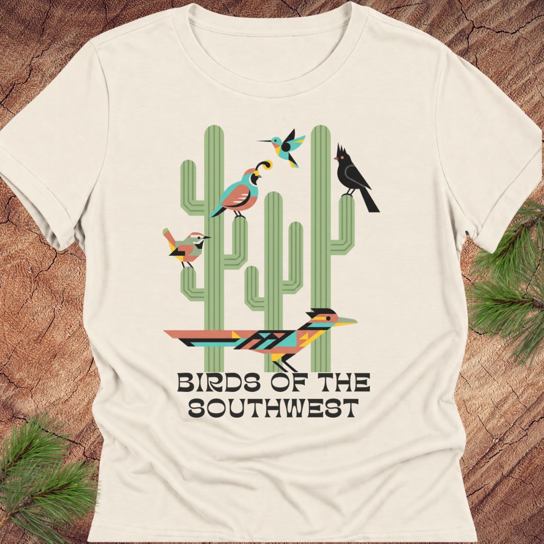 An off-white short-sleeve t-shirt with a colorful graphic of cacti and birds, featuring the text 'BIRDS OF THE SOUTHWEST'. The shirt is displayed flat on a rustic wood background with pine needles in the corners.