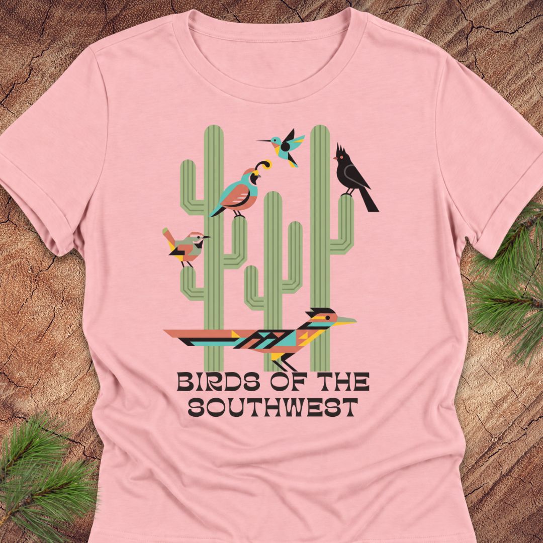 A light pink short-sleeve t-shirt with a colorful graphic of cacti and birds, laid flat on a rustic wood background with pine needles in the corners.