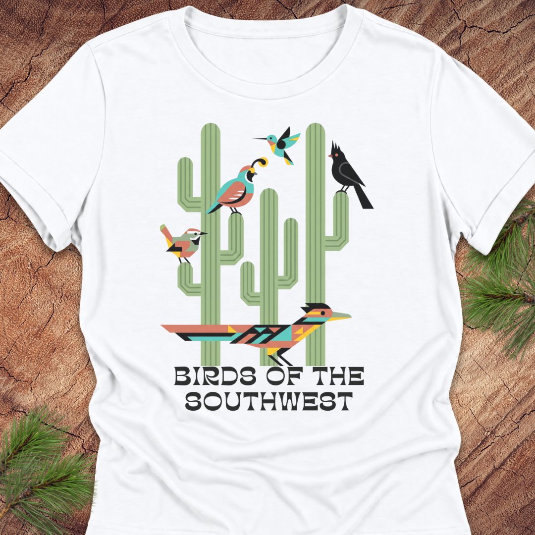 A white short-sleeve crewneck t-shirt featuring a colorful graphic of cacti and birds with the text 'BIRDS OF THE SOUTHWEST', displayed flat on a rustic wood background with pine needles.