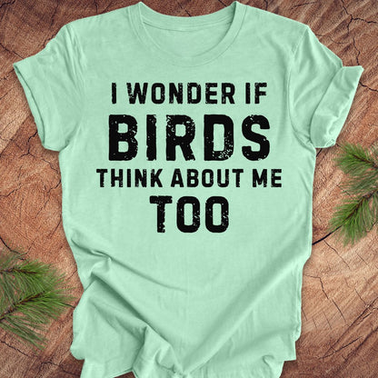 Light green t-shirt with black text 'I wonder if birds think about me too' on a wooden surface.