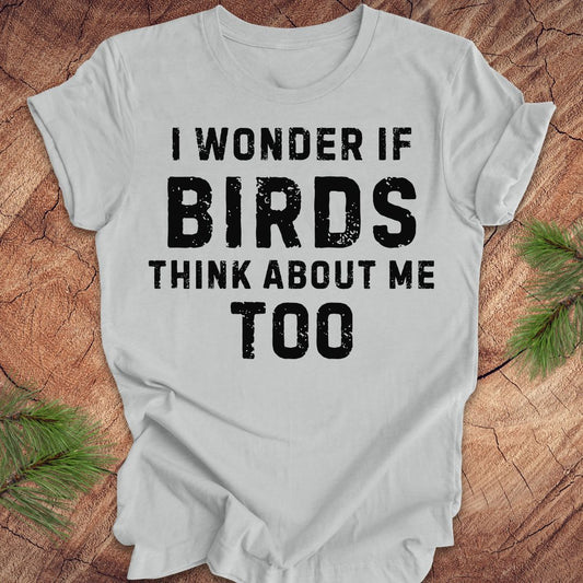 Gray t-shirt with black text 'I wonder if birds think about me too' on a wooden surface with greenery.
