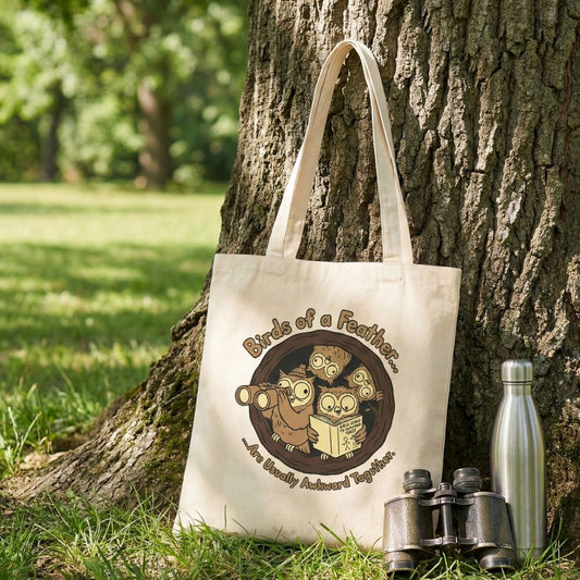 Tote bag with 'Birds of a Feather' design leaning against a tree with binoculars and a water bottle.