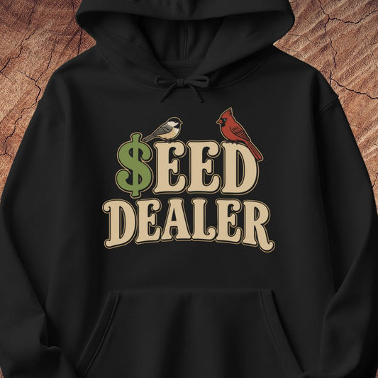 A black pullover hoodie with a graphic print reading 'SEED DEALER' in beige letters, featuring a green dollar sign and two birds (a chickadee and a cardinal) on the chest, displayed against a rustic wood background.