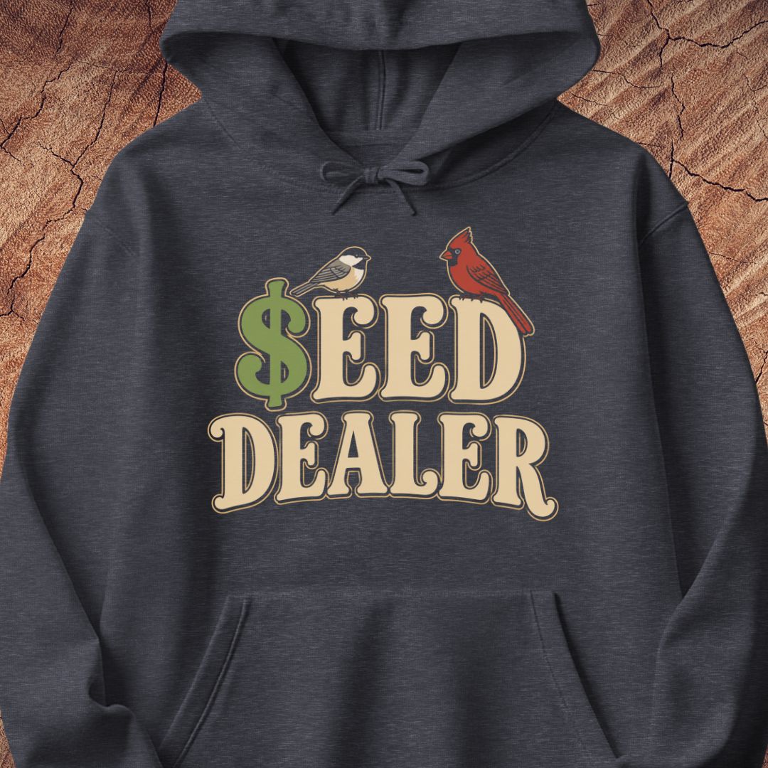 A dark heather gray hoodie featuring a graphic print that reads 'SEED DEALER' in beige varsity-style letters, with a green dollar sign and two birds (a chickadee and a cardinal) on the chest, displayed against a rustic wood background.