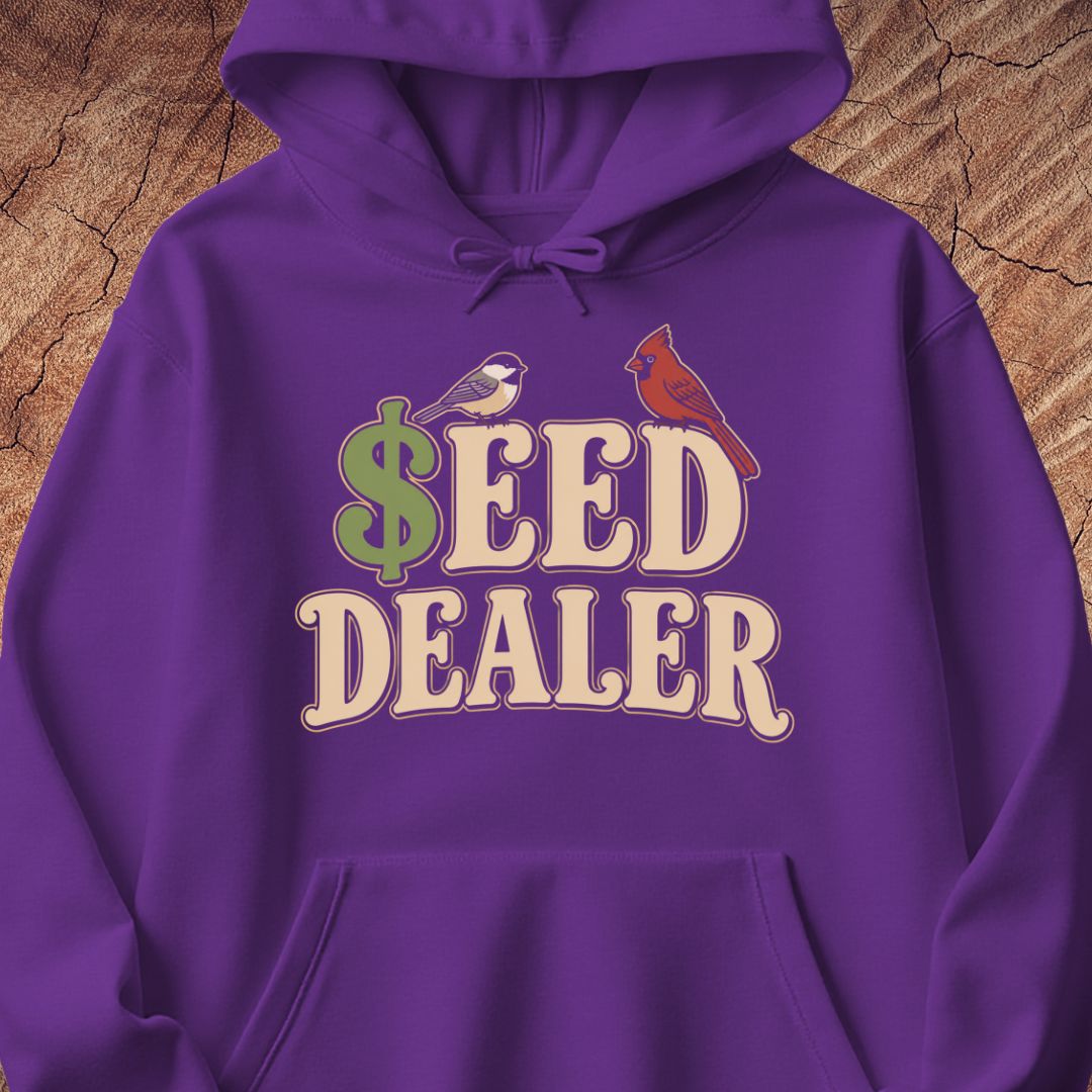 A purple pullover hoodie with a graphic print reading '$EED DEALER' in cream letters, featuring a green dollar sign and two small birds, displayed against a wood texture background.