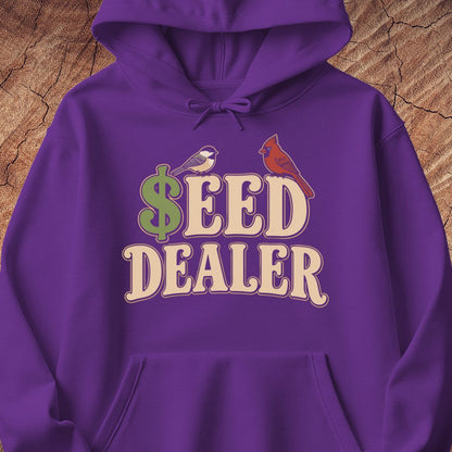 A purple pullover hoodie with a graphic print reading '$EED DEALER' in cream letters, featuring a green dollar sign and two small birds, displayed against a wood texture background.
