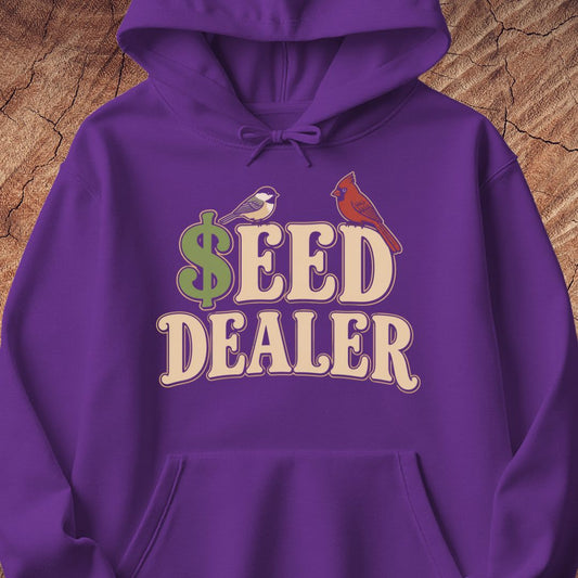 A purple pullover hoodie with a graphic print reading '$EED DEALER' in cream letters, featuring a green dollar sign and two small birds, displayed against a wood texture background.