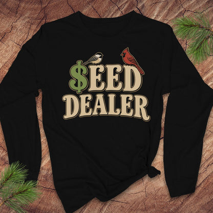 A black long-sleeve t-shirt with a 'SEED DEALER' graphic printed on the chest, styled flat on a rustic wood background with pine branches.