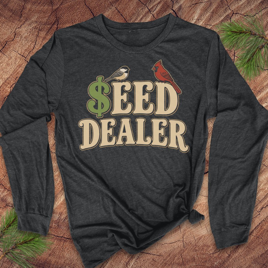 A dark charcoal gray long-sleeve t-shirt with a 'SEED DEALER' graphic print featuring a green dollar sign, a small bird, a cardinal, and beige lettering, laid flat on a rustic wood background with pine branches.