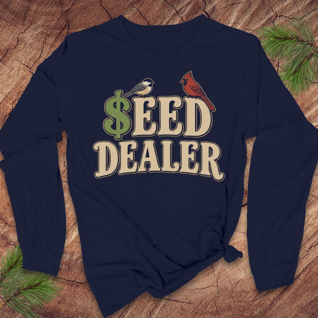 A navy blue long-sleeve t-shirt with a graphic print reading '$EED DEALER' in beige and green, featuring a small bird on the dollar sign and a red cardinal on the E, laid flat on a rustic wood background with pine branches.