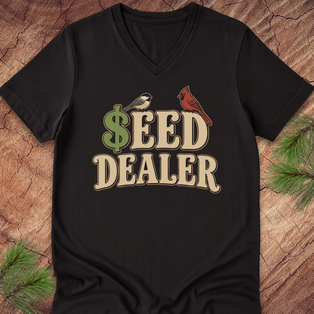 A black V-neck t-shirt laid flat on a rustic wood background with pine branches in the corners. The shirt features a graphic print that reads '$EED DEALER' in beige letters, with a green dollar sign and birds.