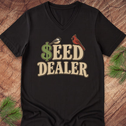 A black V-neck t-shirt laid flat on a rustic wood background with pine branches in the corners. The shirt features a graphic print that reads '$EED DEALER' in beige letters, with a green dollar sign and birds.