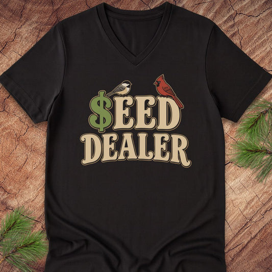 A black V-neck t-shirt laid flat on a rustic wood background with pine branches in the corners. The shirt features a graphic print that reads '$EED DEALER' in beige letters, with a green dollar sign and birds.