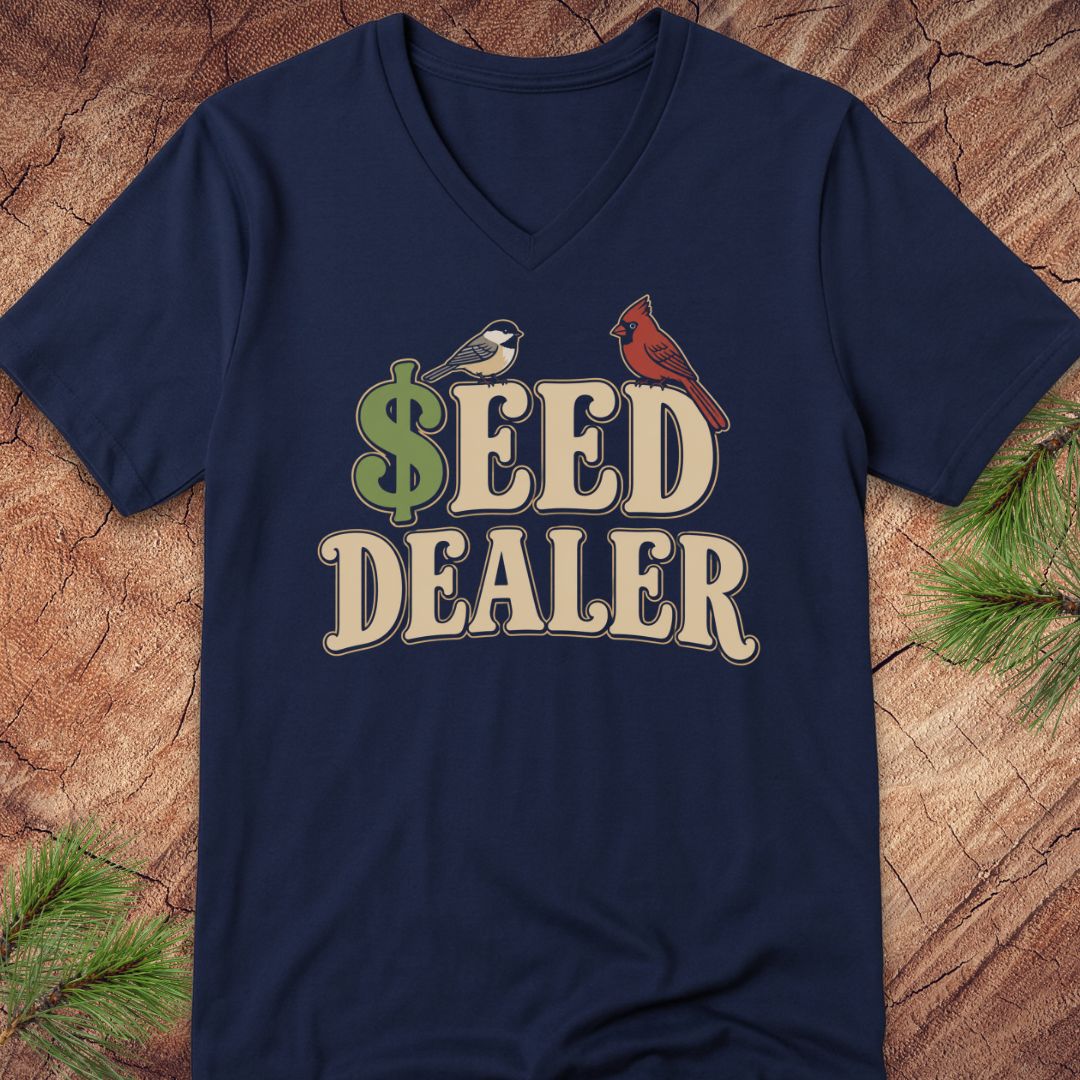 A navy blue V-neck t-shirt with a graphic reading '$EED DEALER' in beige letters, featuring a green dollar sign and two birds (a small white bird and a red cardinal), laid flat on a rustic wood background with pine branches.